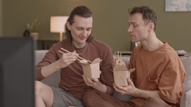romántico caucásico gay pareja sentado en sofá en sala de estar en su apartamento comer china comida de takeout cajas - Imágenes, Vídeo