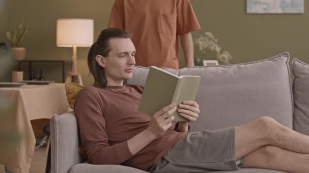 Joven hombre caucásico relajarse en el sofá en la sala de estar con libro, mientras que su novio acariciando su cabello con ternura - Metraje, vídeo