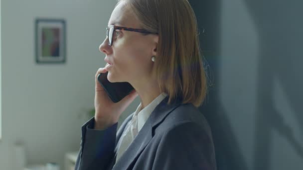 Vista laterale petto up shot di donna d'affari bionda in abbigliamento formale e occhiali che parlano sul cellulare in ufficio - Filmati, video