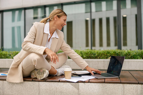 Κομψή γυναίκα διευθυντής εργάζεται με τα έγγραφα και να χρησιμοποιήσετε φορητό υπολογιστή κάθεται έξω από το γραφείο - Φωτογραφία, εικόνα