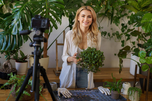 Plantminnende blogger in een wit shirt en spijkerbroek, glimlachend in het natuurlijke licht van haar met planten gevulde stedelijke appartement - Foto, afbeelding