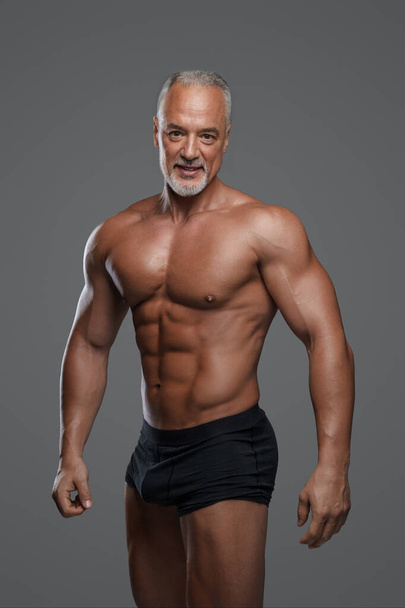 Ελκυστικός ηλικιωμένος άνδρας με μυϊκή σωματική διάπλαση, κομψή γκρίζα γενειάδα, μαύρα εσώρουχα και γυμνό κορμό, ποζάροντας σε γκρι φόντο - Φωτογραφία, εικόνα