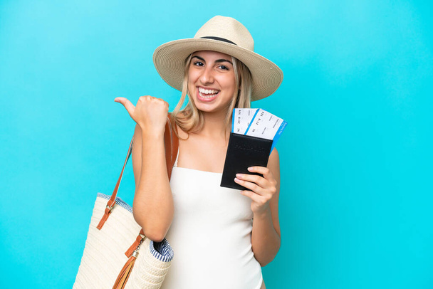 Молода біла жінка в купальнику тримає паспорт ізольовано на синьому фоні, вказуючи на бік, щоб представити продукт
 - Фото, зображення