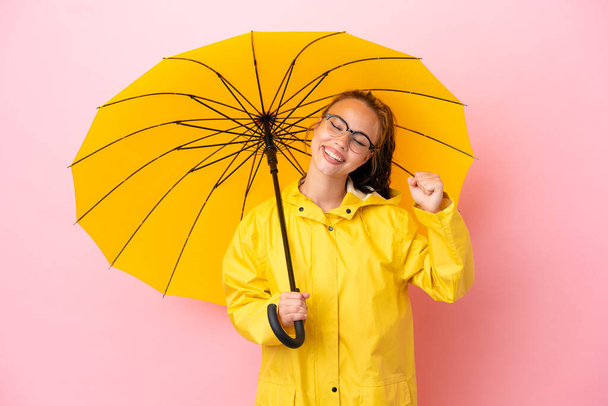 Yağmura dayanıklı paltosu ve şemsiyesi olan genç Rus kız pembe arka planda zaferi kutluyor. - Fotoğraf, Görsel