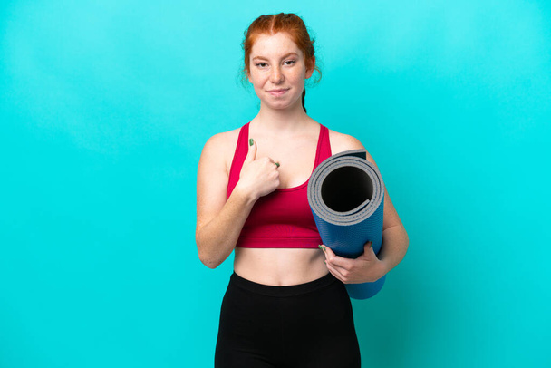 Jeune femme rougeâtre sportive allant à des cours de yoga tout en tenant un tapis isolé sur fond bleu avec expression faciale surprise - Photo, image