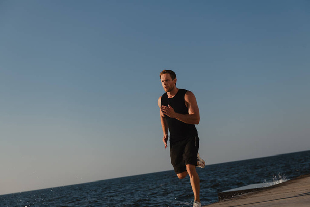 Confident athletic man in sports clothing running outdoors with the sea on background - Photo, Image
