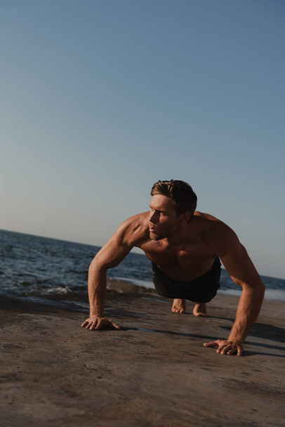 Handsome young shirtless man doing push-ups while exercising outdoors with the sea on background - Photo, Image