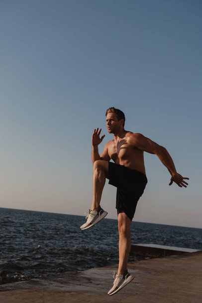 Full length of strong young shirtless man jumping outdoors with the sea on background - Foto, imagen