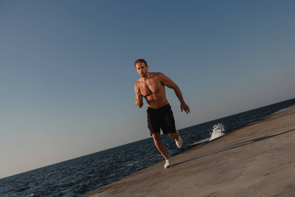 Confident athletic man with muscular torso running outdoors with the sea on background - Photo, Image