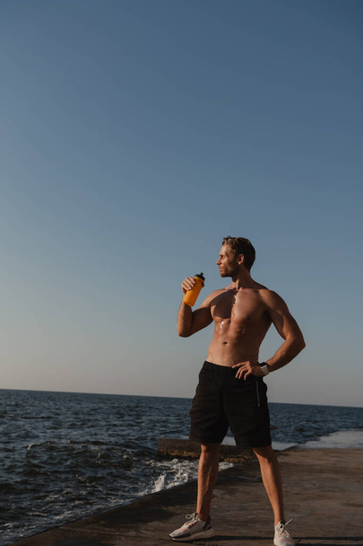 Handsome male athlete drinking water while relaxing after sport training seaside - Photo, Image