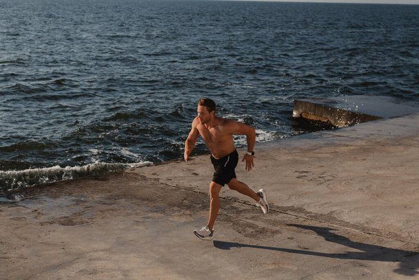 Top view of confident athletic man jogging by the pier with the sea on background - Photo, Image