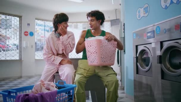 Teenage couple talking together in public laundromat. Hipster girl guy washing clothes waiting in self service. Muscular hispanic teen holding basket enjoy communication. Carefree high school friends - Footage, Video