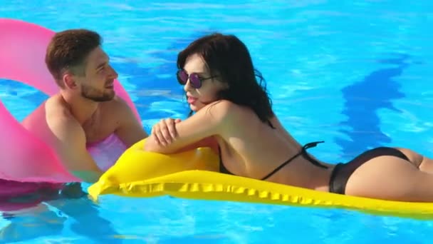 A couple of young lovers talking in the pool. Medium shot: a man and a woman enjoy relaxing outdoors in the pool on inflatable rings and a swimming mattress. - Footage, Video