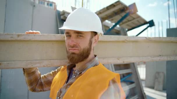Pohled na pracovníka s dlouhým těžkým trámem. Mužský pohled do kamery. Zarámovat stavbu. Slunečný den - Záběry, video