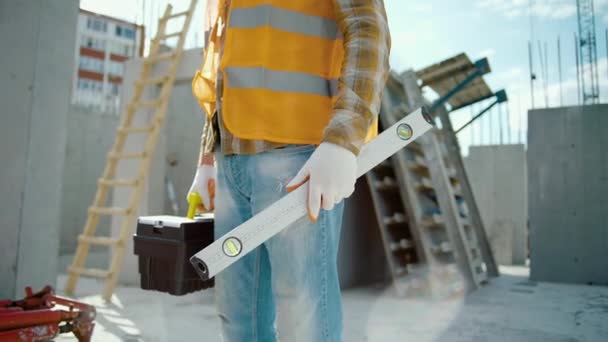 Trabajador de construcción con medidor de nivel de burbuja y caja de herramientas de pie en el sitio de construcción - Metraje, vídeo