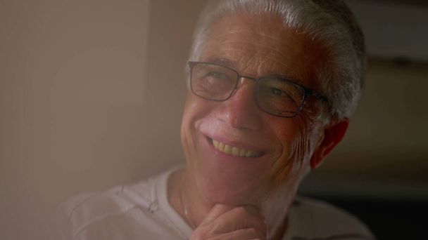 Κοντινό πλάνο ενός χαρούμενου ηλικιωμένου άνδρα με ώριμη έκφραση που χαμογελά κοιτάζοντας έξω από το παράθυρο - Φωτογραφία, εικόνα