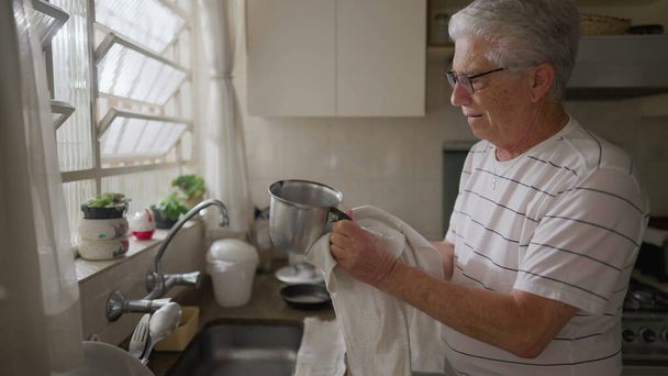Gelukkige oudere man die afwas bij de gootsteen droogt. Authentieke huiselijke scene levensstijl van volwassen oudere persoon thuis ritueel - Foto, afbeelding