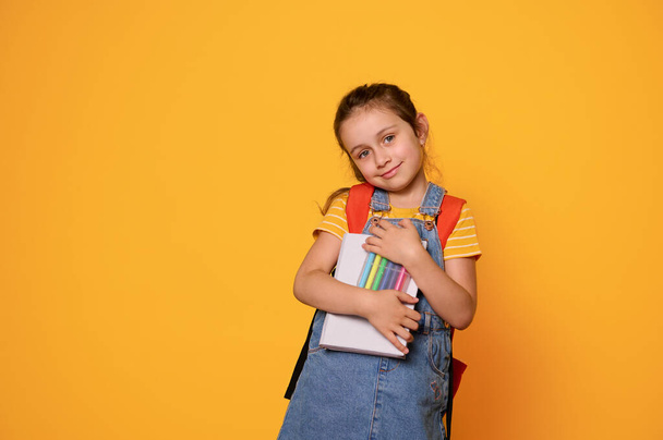 Красивая маленькая девочка с войлочными ручками, с рюкзаком, мило улыбается, смотрит в камеру, изолированная на фоне оранжевой студии. Готовлюсь к новому учебному году. Дети и концепция образования - Фото, изображение