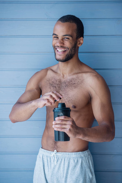 Όμορφος αθλητής κρατά ένα σέικερ με πρωτεΐνη και χαμογελά ενώ στέκεται τόπλες μπροστά από γκρίζο τοίχο - Φωτογραφία, εικόνα