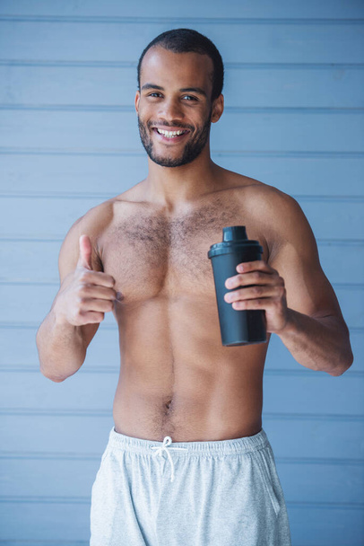 Esportista bonito está segurando uma coqueteleira com proteína, mostrando Ok sinal e sorrindo enquanto em pé topless na frente da parede cinza - Foto, Imagem