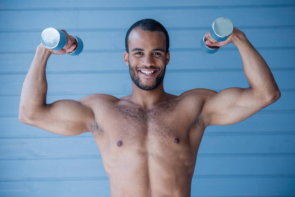 Esportista bonito está segurando halteres, olhando para a câmera e sorrindo enquanto está de pé topless na frente da parede cinza - Foto, Imagem