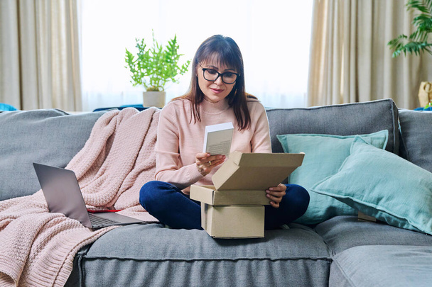 Mature woman sitting on sofa at home, living room, unpacking cardboard box with online shopping, buying new mobile phone smartphone. Internet shopping, postal delivery, delivery service - Foto, Imagen