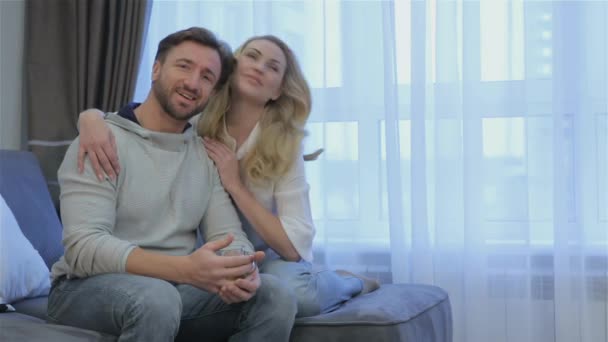 Nice couple sitting on the couch at home. Pretty blond woman sitting behind brunette bearded man. Casual home portrait of attractive couple - Footage, Video