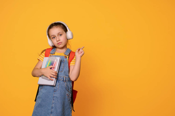 ヘッドフォンとカジュアルなデニムオーバーオールのスマートな1年生の女子は,コピースペースの脇にポイント,広告テキストのためのモックアップ,孤立したオレンジスタジオの背景. 学校に戻る。 教育について。 子供たち - 写真・画像