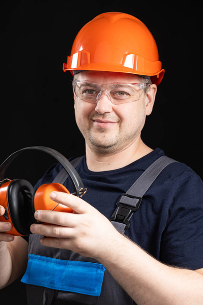 Οικοδόμος σε ένα προστατευτικό κράνος με ακουστικά ακύρωσης θορύβου στα χέρια του σε μαύρο φόντο. - Φωτογραφία, εικόνα