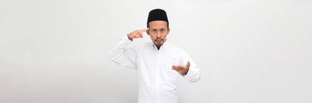 An angry young Indonesian man wearing a songkok or peci or kopiah gestures with his finger against his temple, asking Are you crazy? He is isolated on a white background - Photo, Image