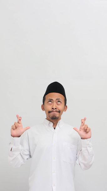 Возбужденный молодой индонезиец, одетый в сонгкок, печи или копию, делает скрещенный жест и смотрит вверх на пустое копировальное пространство, изолированное на белом фоне - Фото, изображение