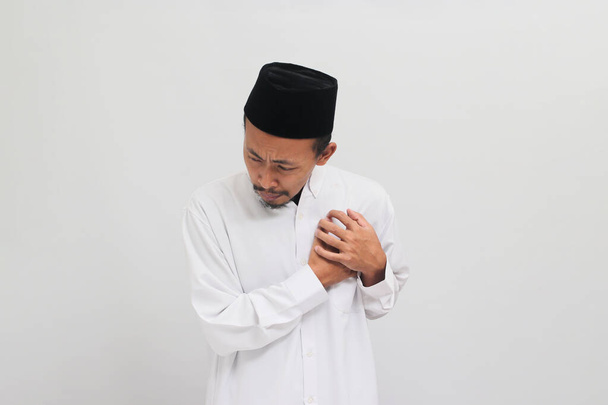Jonge Indonesische man, het dragen van een songkok, peci, of kopiah, grijpt zijn borst, het ervaren van acute pijn, die een symptoom van een hartaanval kan zijn, geïsoleerd op witte achtergrond - Foto, afbeelding