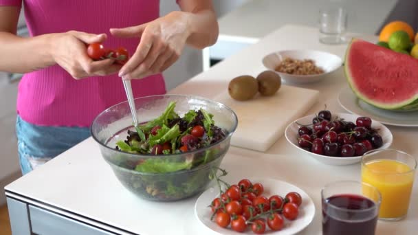 Langzame beweging van een onherkenbare vrouw aan de keukentafel en het gieten van kerstomaten in een kom met salade en bladgroenten. - Video