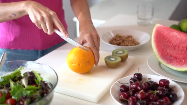 Movimento lento da colheita fêmea cortando laranja a bordo na mesa contra salada e cerejas com melancia e, em seguida, mostrando metades - Filmagem, Vídeo