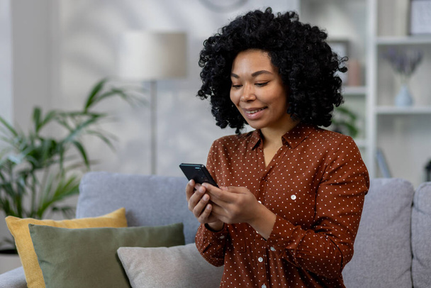 Nuori kaunis Afrikkalainen Amerikkalainen nainen istuu sohvalla kotona, nainen kihara tukka tilalla puhelin käsissä, käyttäen online-sovellus olohuoneessa selailu Internet-sivuja ja kirjoittamalla viestejä - Valokuva, kuva