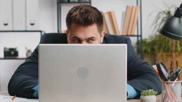 Впевнений молодий бізнесмен ховається за ноутбуком, дивлячись на камеру, шпигуючи за своїми колегами, працюючи, підглядаючи. Професійний фрілансер, дивлячись з-за комп'ютера з хитрим поглядом на очі - Кадри, відео