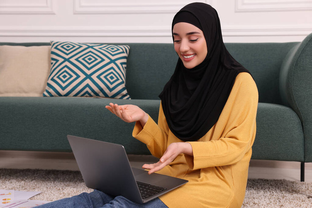 Moslim vrouw in hijab met behulp van video chat op laptop in de buurt van bank binnen - Foto, afbeelding