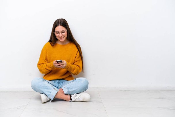 Chica joven sentada en el suelo aislada sobre fondo blanco enviando un mensaje con el móvil - Foto, imagen