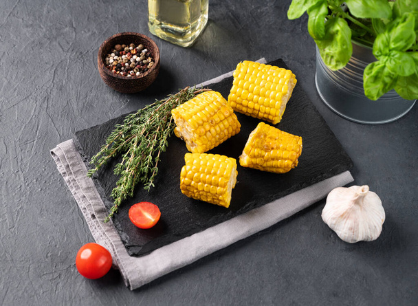 Grilled sweet corn on a black slate on a dark background with fresh vegetables and herbs. Healthy vegetarian food concept. Top view. - Photo, Image