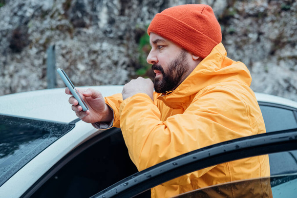 Молодий бородатий чоловік використовує телефон, зупиняючись у дорожній подорожі. Він носить повсякденну жовту куртку і червоний капелюх. Осінні подорожі на машині. - Фото, зображення