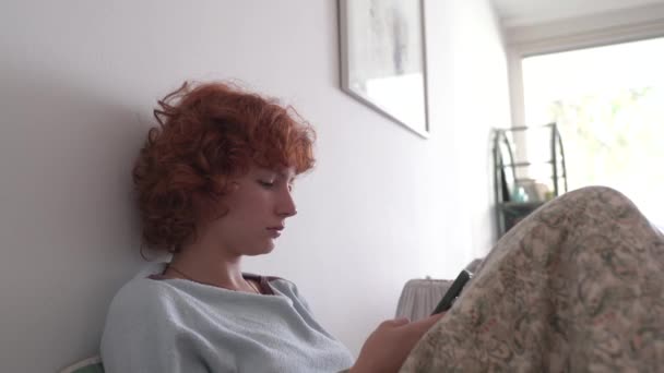 Chica adolescente pelirroja rizada, en pijama, se sienta en la cama, utiliza móviles, muecas, escribe mensaje en línea - Imágenes, Vídeo