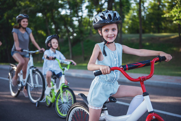 Onnellinen perhe ajaa pyörillä ulkona ja hymyilee. Pieni tyttö etualalla katsoo kameraa.
 - Valokuva, kuva