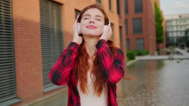 Békés lány modern vezeték nélküli fejhallgatóval zenét hallgat az utcán, vörös hajú nő alkalmi piros sima pólóban..  - Felvétel, videó