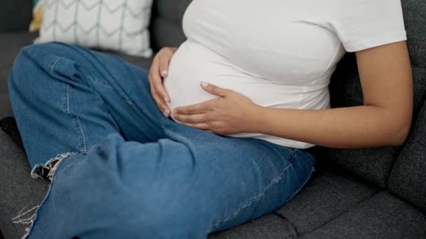 Молодая беременная женщина трогает живот, сидя дома на диване. - Кадры, видео