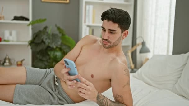 Joven hispano haciendo videollamada sentado en la cama en el dormitorio - Imágenes, Vídeo