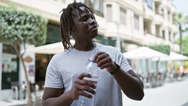 Afroamerikaner trinkt lächelnd Wasserflasche auf Café-Terrasse - Filmmaterial, Video