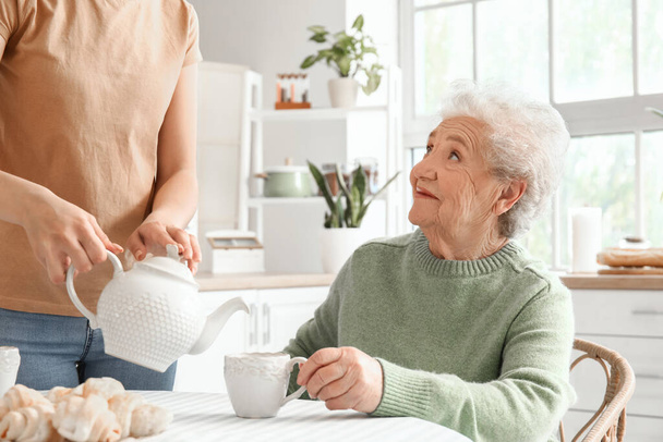 Egy fiatal nő teát tölt csészébe a nagyanyjának a konyhában. - Fotó, kép