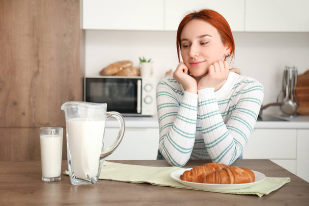 Fiatal vörös hajú nő egy pohár tejjel és croissant a konyhában - Fotó, kép