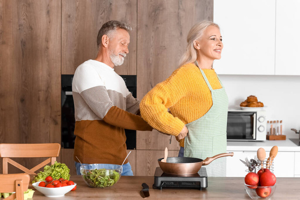 volwassen man koppelverkoop schort op zijn vrouw in keuken - Foto, afbeelding