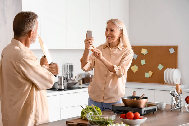 Mature woman taking picture of her husband with knives in kitchen - Valokuva, kuva